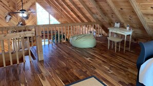 Une chambre mansardée avec des poutres apparentes, un ventilateur de plafond, un fauteuil poire, une petite table et une chaise.
