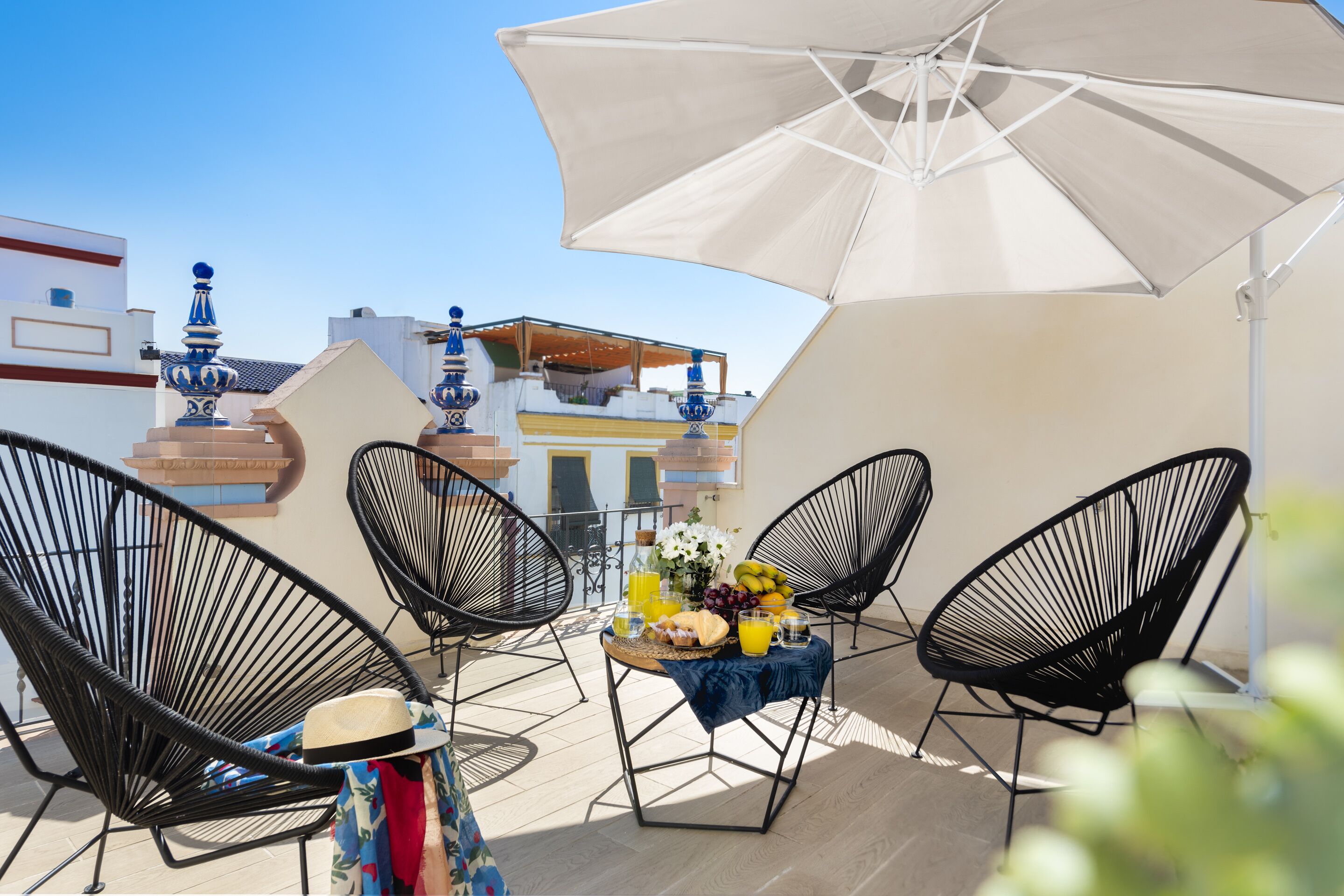 Una terraza con una mesa preparada para dos personas, dos sillas negras de alambre, un paraguas blanco y una mesa con frutas y bebidas.