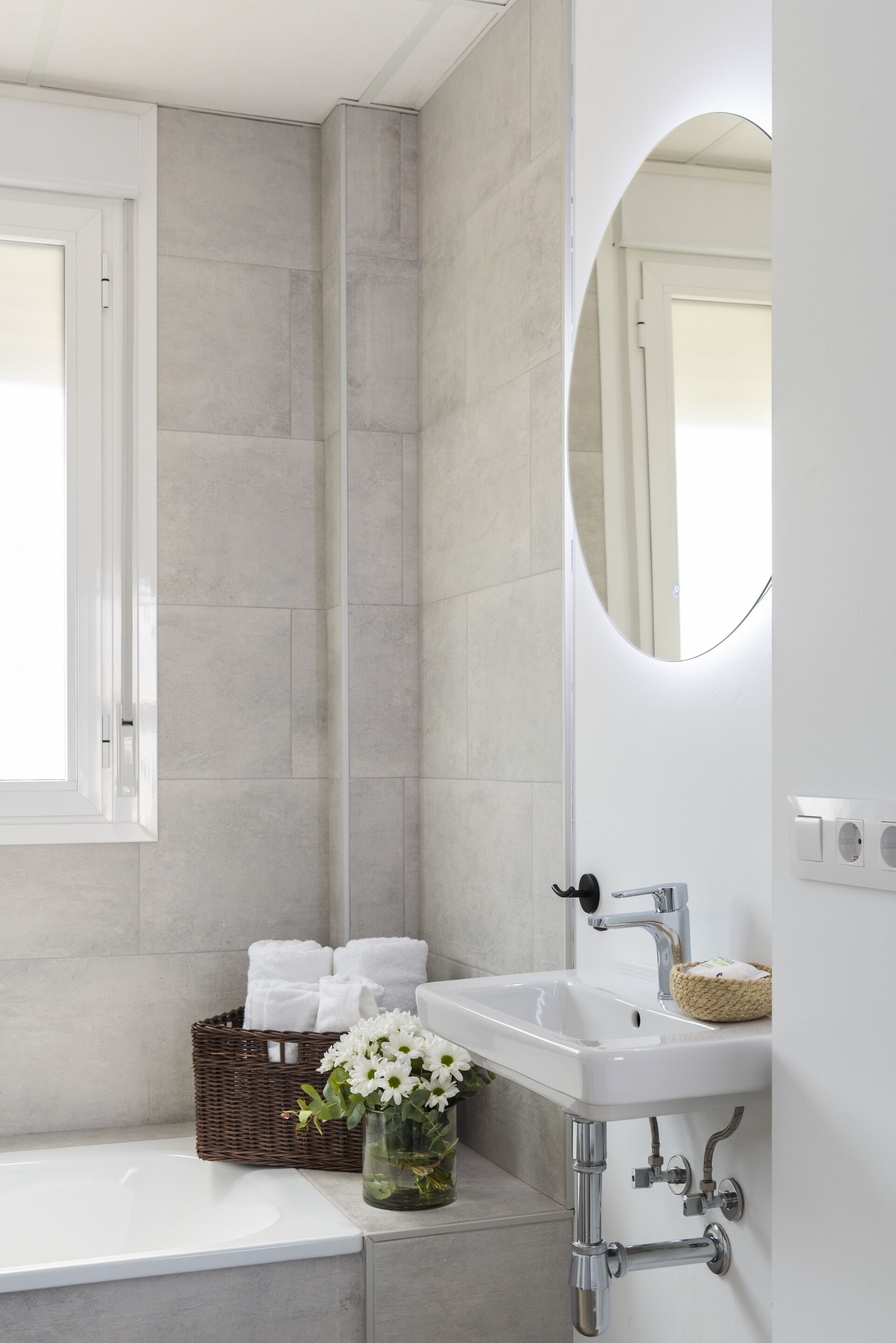 Un baño moderno con un lavamanos blanco, un espejo, una bañera y una cesta de mimbre con toallas.