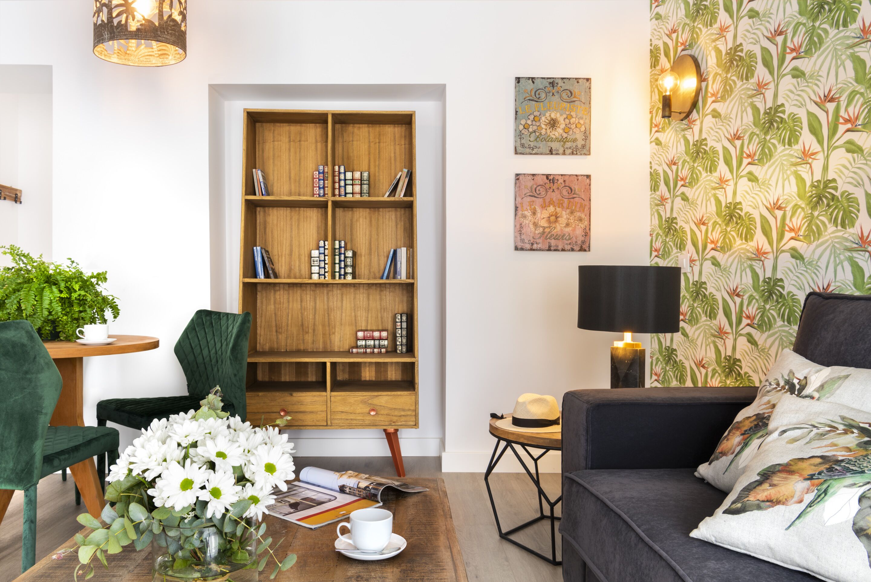 En hyggelig stue med en træbogreol, en grøn lænestol, en mørk sofa og et sofabord med en vase blomster.