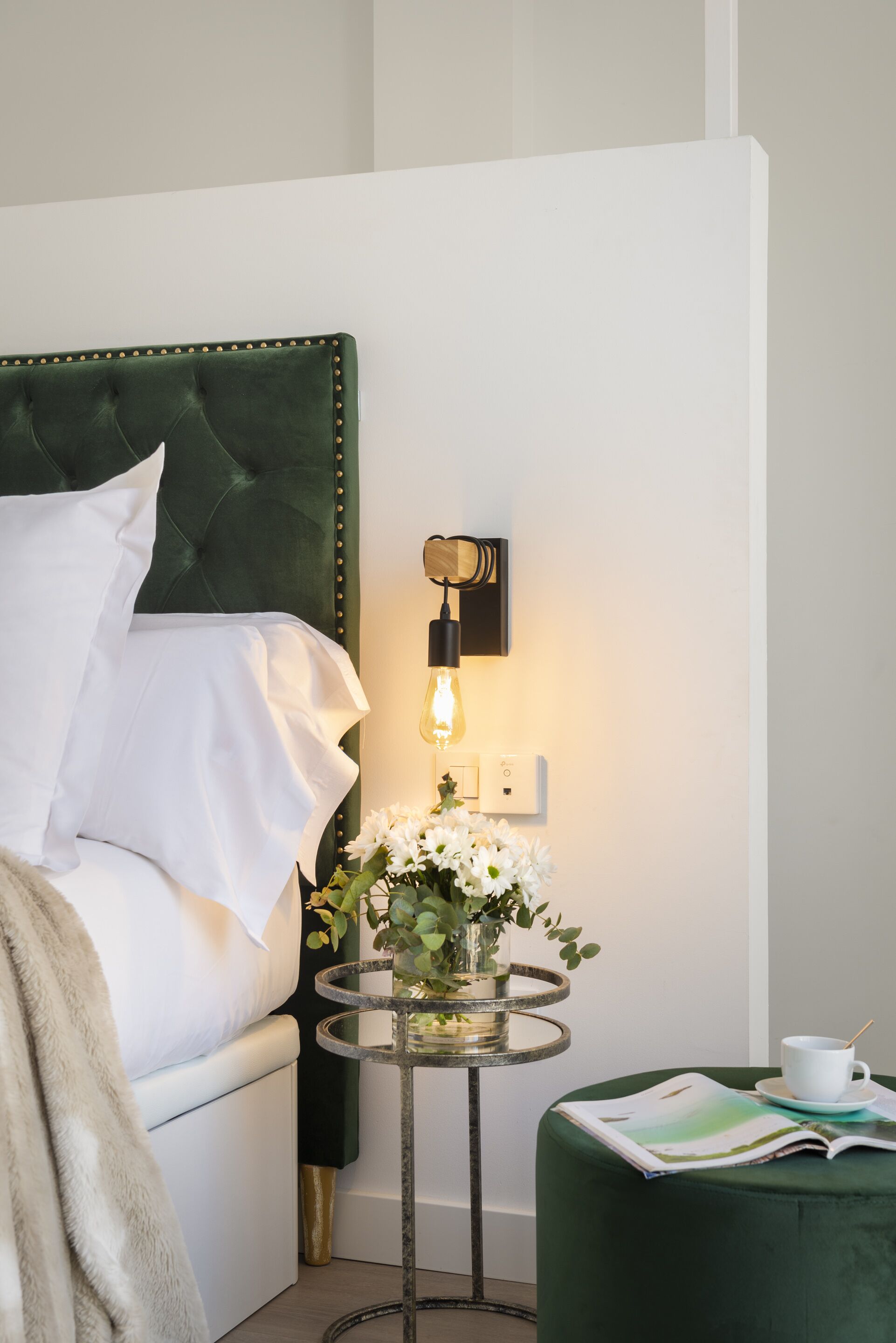 Una cama bien tendida con sábanas blancas, un cabecero tapizado en verde, una mesita de noche con un jarrón de flores y una pequeña mesa redonda con una taza y una revista.
