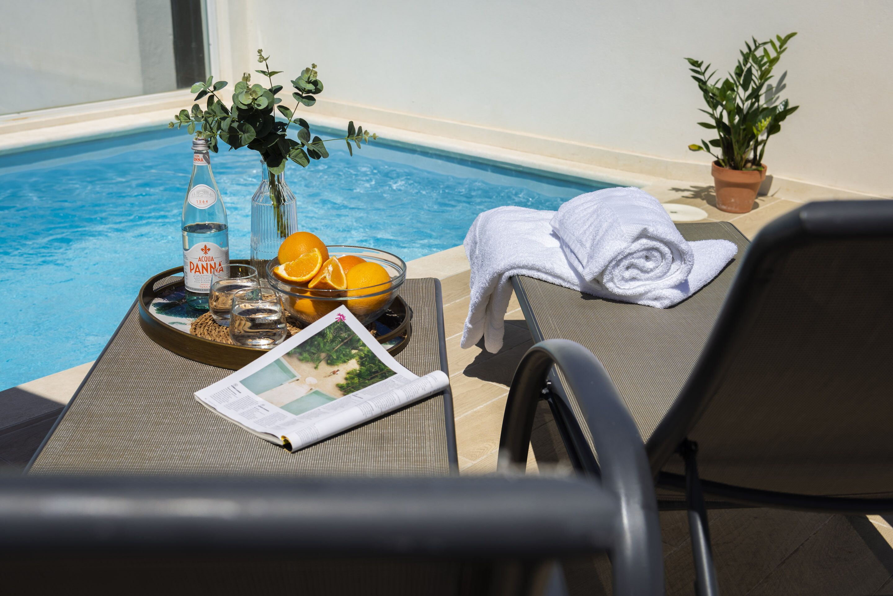 Zona junto a la piscina con una bandeja que contiene una botella de agua, vasos, naranjas y una revista.