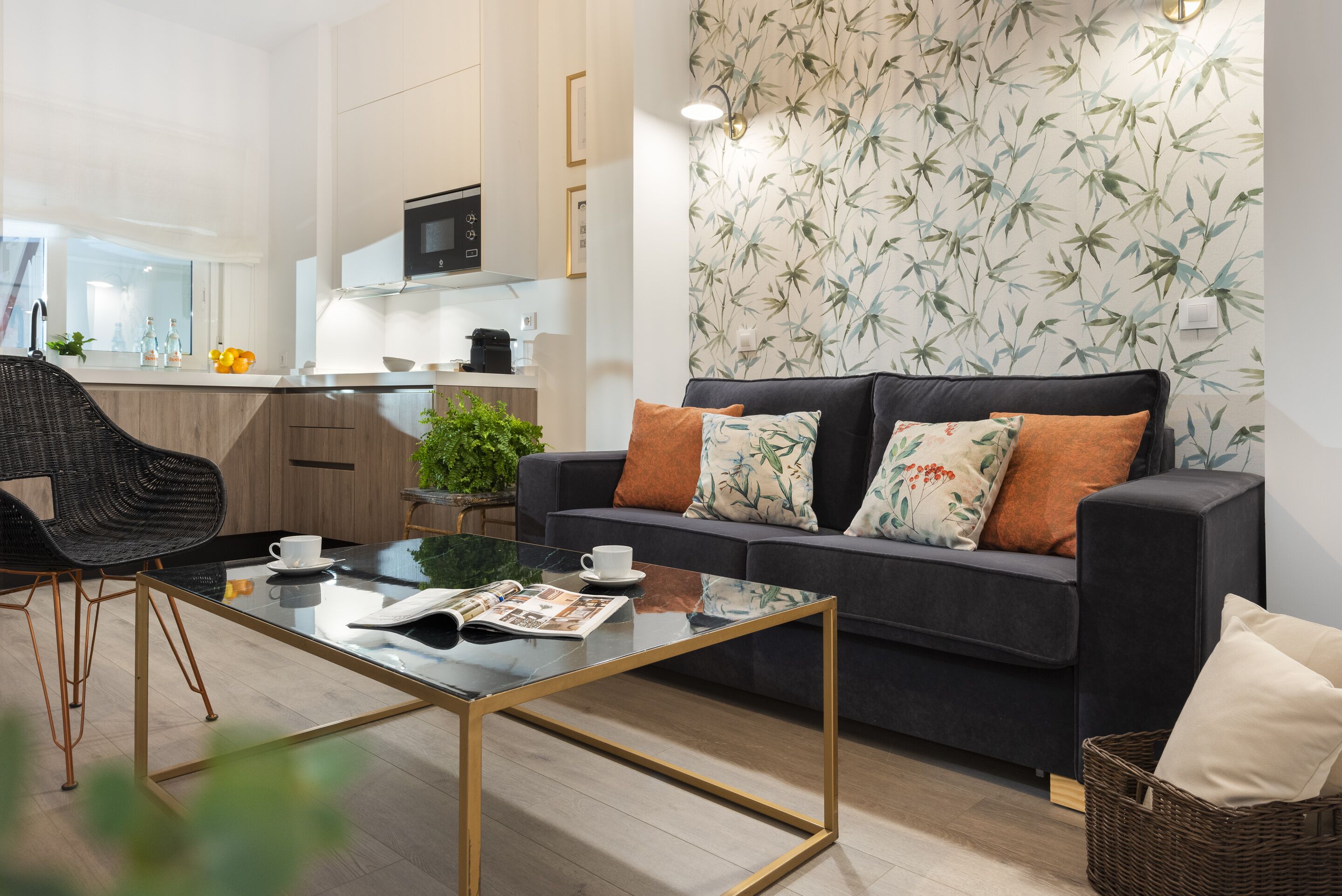 Een moderne woonkamer met een donkere bank, een glazen salontafel en een keuken op de achtergrond.