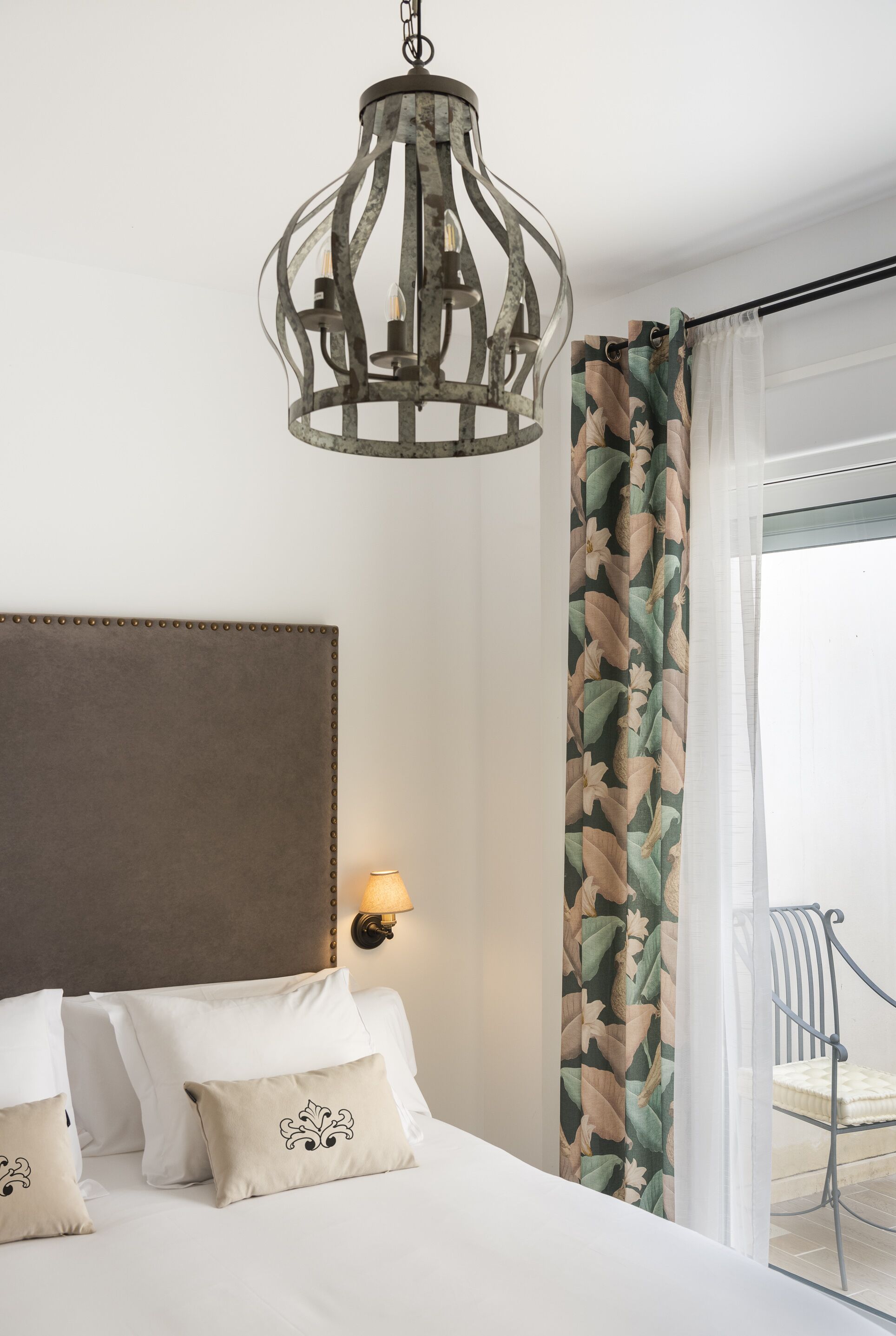 Habitación de hotel con una cama grande, una lámpara colgante decorativa, una lámpara de pared y vistas al exterior a través de una ventana con cortinas translúcidas.