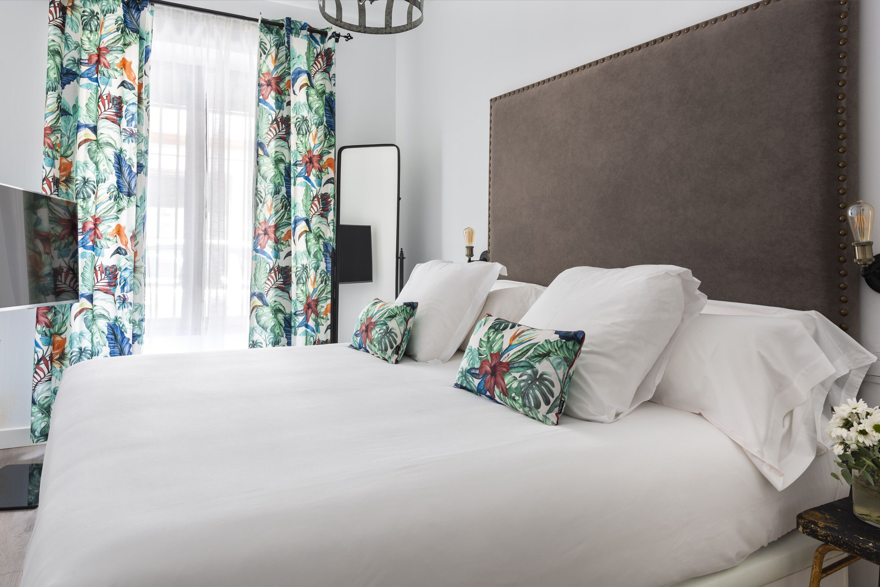 A neatly made bed with white linens and tropical patterned pillows, a large mirror, and a modern headboard in a well-lit room.