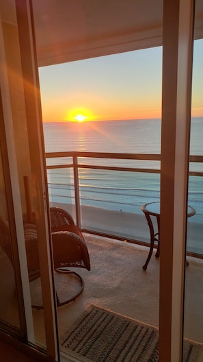 Uma varanda com vista para o pôr do sol sobre o oceano, uma cadeira de vime e uma mesa pequena.