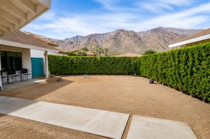 A courtyard with a view of mountains, a neatly trimmed hedge, and a patio area with chairs.