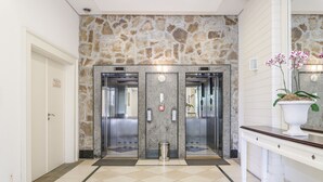 Un vestíbulo con dos ascensores, una pared de piedra, una puerta blanca y una zona de recepción con una planta.