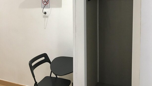 A room with a desk, chair, and a wardrobe with hanging clothes.