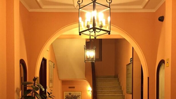 A grand staircase with a chandelier, leading to a landing with a framed picture and potted plants. - LM Rooms Opera (Madrid)