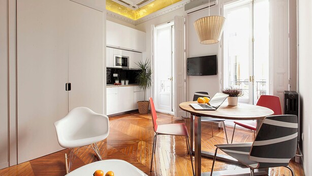 Ein modernes Wohnzimmer mit Essbereich, mit Holzboden, rundem Tisch mit Stühlen, Fernseher an der Wand und einer Küche im Hintergrund.
