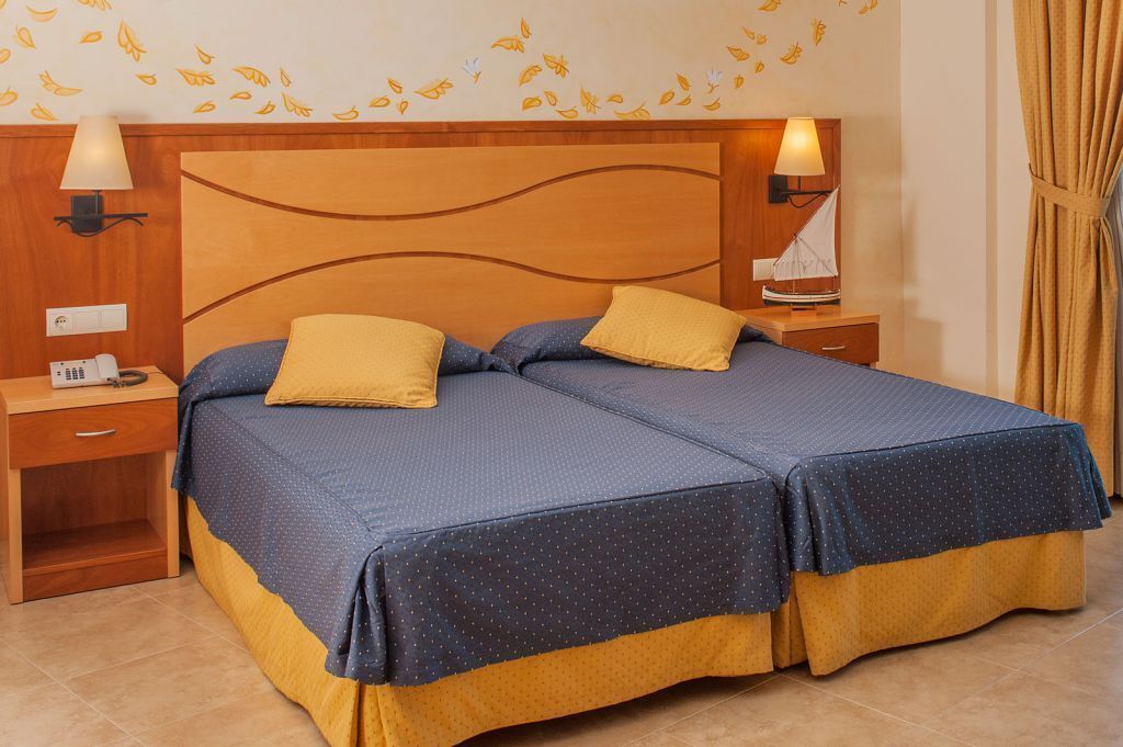 Cama doble con dos almohadas, cabecera de madera, lámparas de noche, mesita de noche y pared decorada con motivos de hojas.