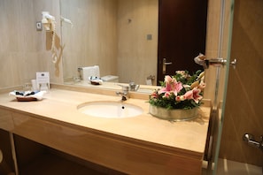 A hotel bathroom with a large sink, a mirror, a hairdryer, and a flower arrangement.
