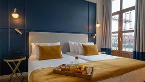 Habitación de hotel con una cama grande, dos mesitas de noche, una bandeja con alimentos para el desayuno y vista a un edificio a través de la ventana.