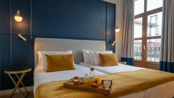 Habitación de hotel con una cama grande, dos mesitas de noche, una bandeja con alimentos para el desayuno y vista a un edificio a través de la ventana.