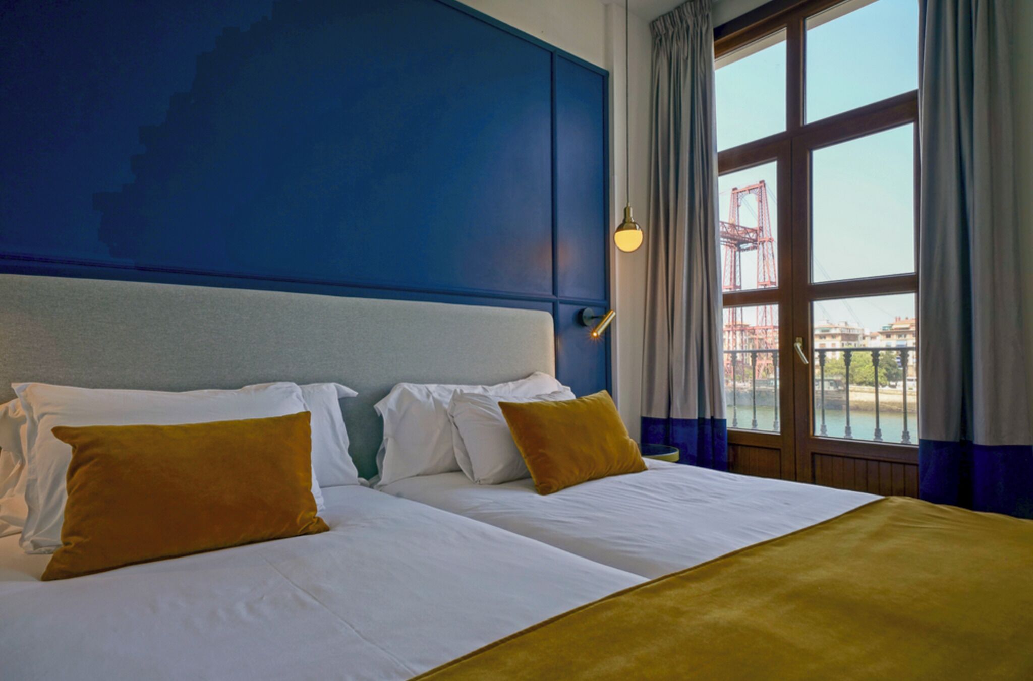 Habitación de hotel con una cama grande, dos almohadas, cabecero, ventana con cortinas y vista a un puente.