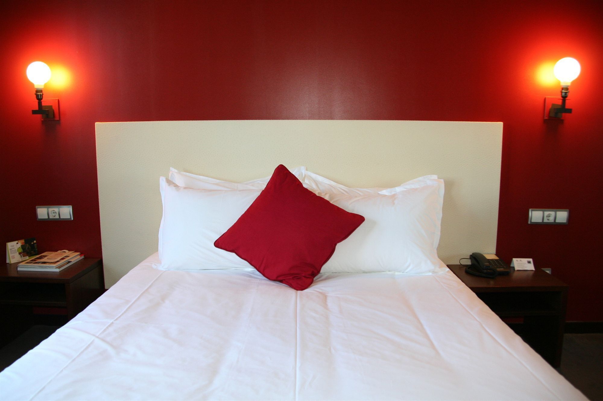 Una cama bien hecha con ropa blanca y una almohada roja, dos mesitas de noche con lámparas, y una pared roja.
