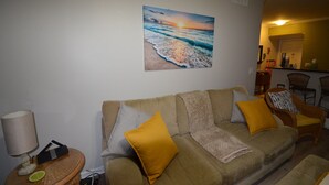 A living room with a beige sofa, yellow and gray cushions, a small round table with a lamp, and a wall-mounted painting of a beach scene. - Exceptional Luxury Resort (Boynton Beach)