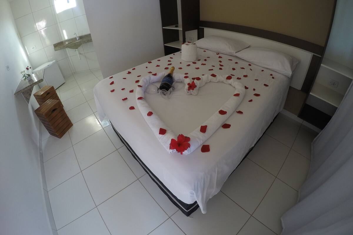 Una habitación de hotel con una cama bien hecha, una mesita de noche con una botella y vasos, y un taburete de madera.