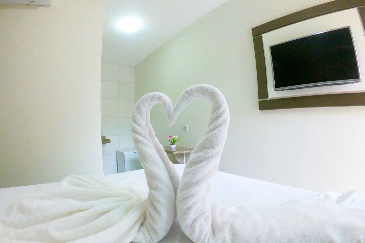 Una habitación de hotel con cama, una toalla dispuesta en forma de corazón, un televisor montado en la pared y una pequeña planta sobre una mesa.