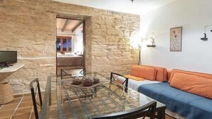 Un salon chaleureux avec un mur en pierre, une table basse en verre, un canapé bleu et un téléviseur posé sur un meuble.
