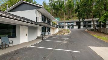 Un motel de dos pisos con estacionamiento, mobiliario exterior y vista a un área boscosa.