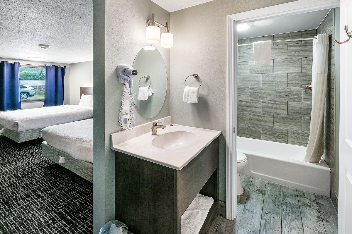 A hotel bathroom with a sink, mirror, and shower. There are two beds in the adjacent room.