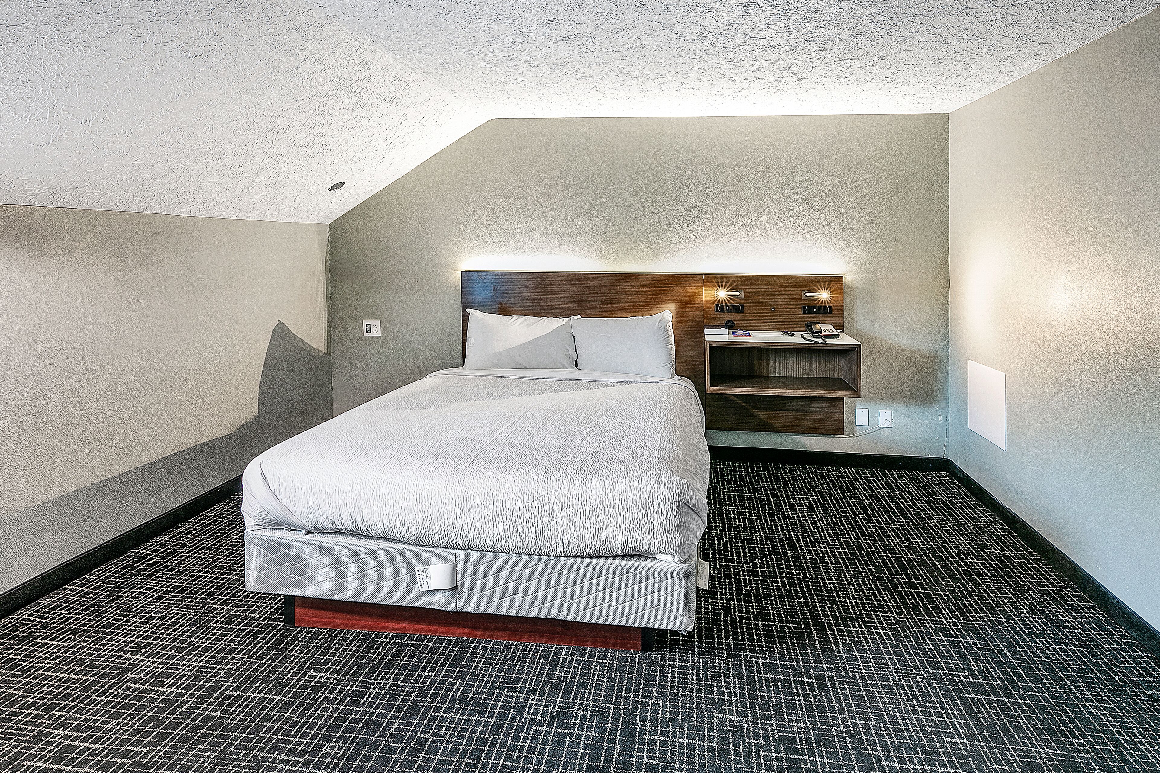 A hotel room with a bed, a nightstand, a wall-mounted lamp, and a small table with a phone and a lamp.