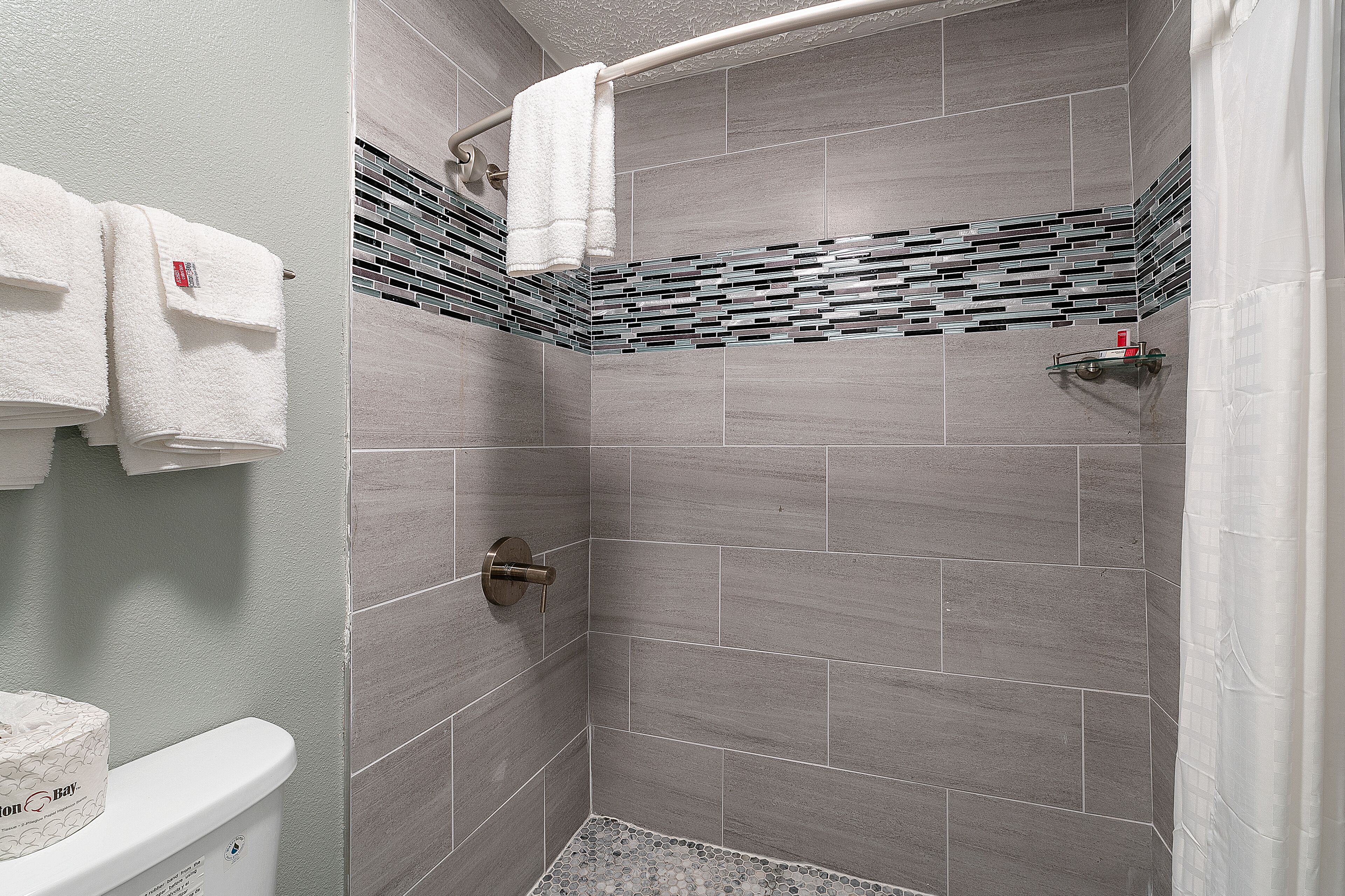 Un baño moderno con azulejos grises, una ducha con repisa de vidrio y un inodoro con portarrollos.