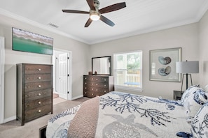 Une chambre à coucher comprenant un lit, une commode, un ventilateur de plafond, un tableau, une fenêtre avec des stores et un tableau encadré accroché au mur.