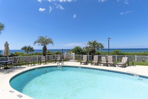 Une piscine d’un bleu éclatant, bordée de chaises longues et d’ombrelles, entourée d’une clôture blanche, et offrant une vue sur l’océan.