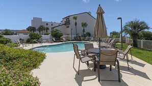 A pool area with lounge chairs and a table, surrounded by greenery and a building in the background. - Grand Playa, Unit 402 | Gulf - Front Condo | Priva (Santa Rosa Beach)