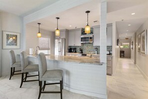 Una cucina moderna con isola centrale, sgabelli da bar, illuminazione a sospensione e vista sull'adiacente sala da pranzo.