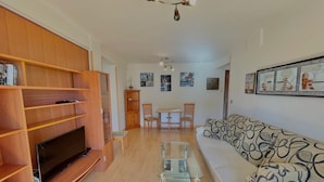 Ein Wohnzimmer mit Fernseher, Holzregalen, einem Glastisch, einem gemusterten Sofa und einem Essbereich mit Stühlen und Tisch.