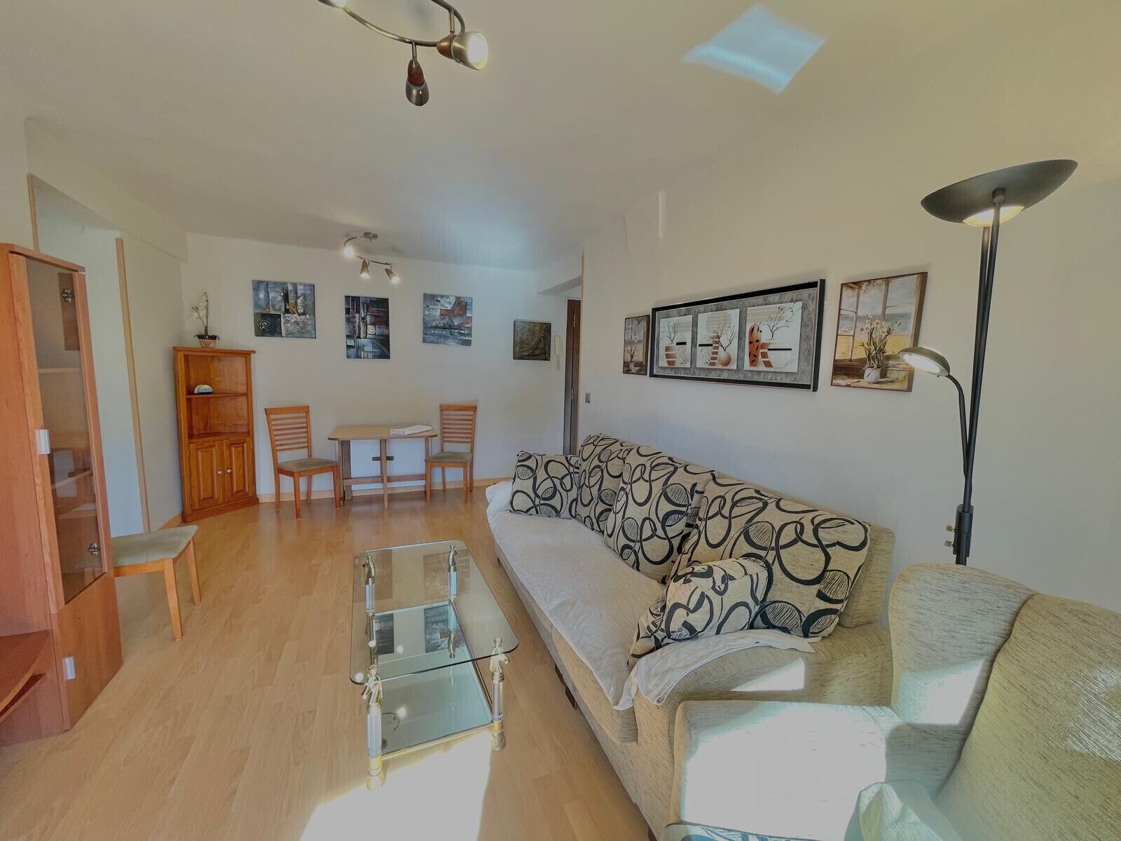 A living room with a sofa, chairs, a glass coffee table, a wooden cabinet, and artwork on the walls.