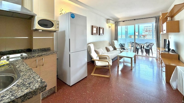 A compact kitchen with a microwave, refrigerator, and granite countertop. There is a seating area with a sofa and chairs, and a dining table. The room has a large window offering a view of the city. - TERRASOL PLAZAMAR 2-6 (Torre del Mar)