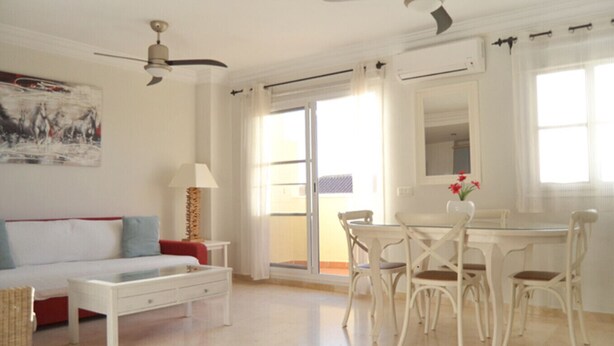 Un salón luminoso con un sofá blanco, una mesita y un comedor con sillas blancas. Hay ventiladores de techo y un ventanal grande que proporciona luz natural.