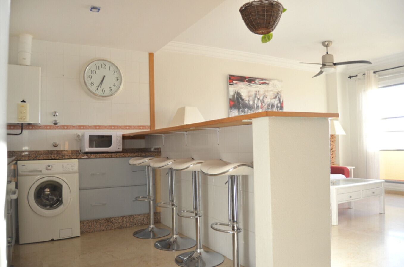 A kitchen with a washing machine, a microwave, and a clock on the wall. There are three bar stools and a ceiling fan.