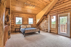 Un dormitorio tipo cabaña de madera con una cama, dos ventanas, una puerta y una escalera.