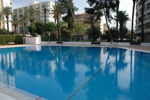 Une piscine à l’eau d’un bleu cristallin, entourée de palmiers et de bâtiments modernes.
