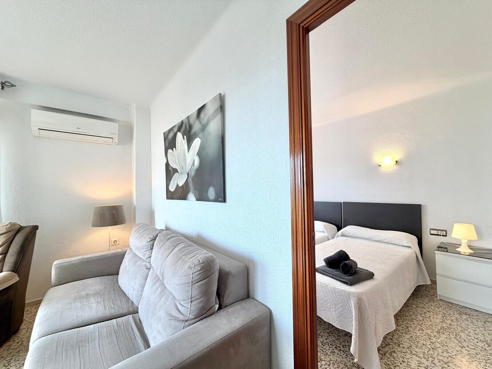 A hotel room with a bed, a sofa, a wall-mounted air conditioner, a painting, and a bedside lamp.