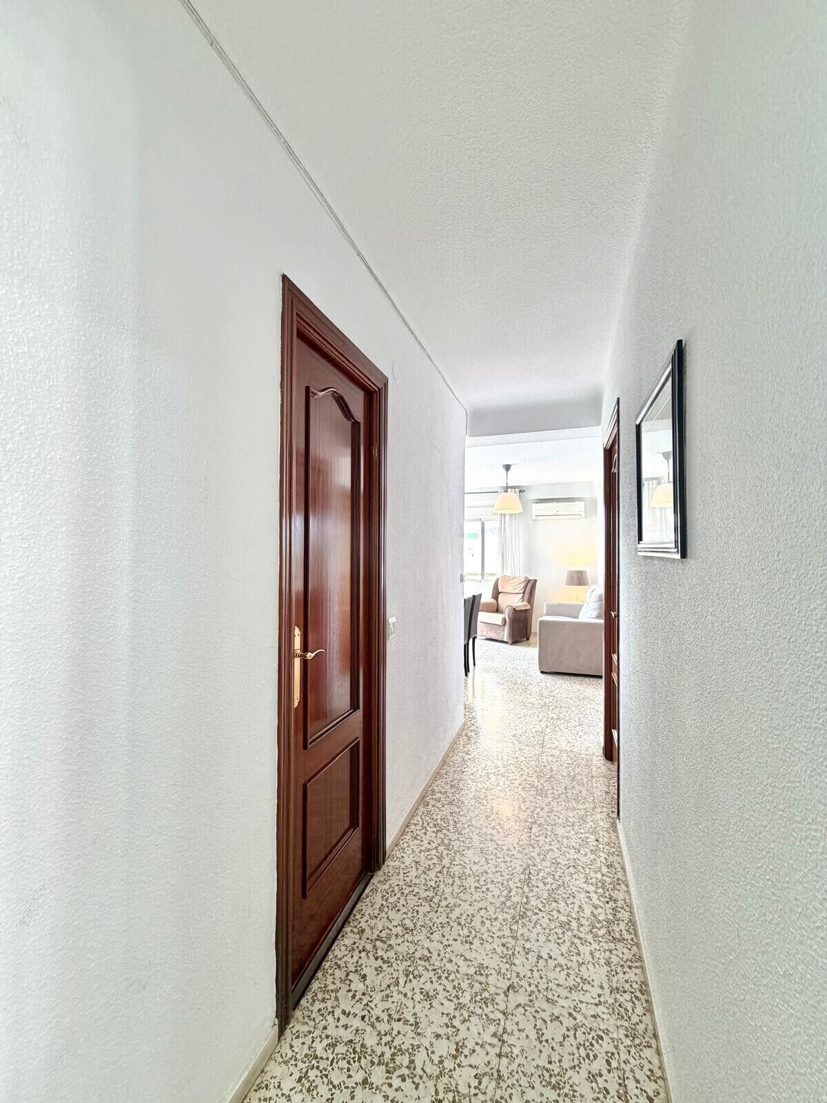 Un pasillo con una puerta de madera, un cuadro en la pared y una vista parcial de una sala de estar con un sofá y una lámpara.