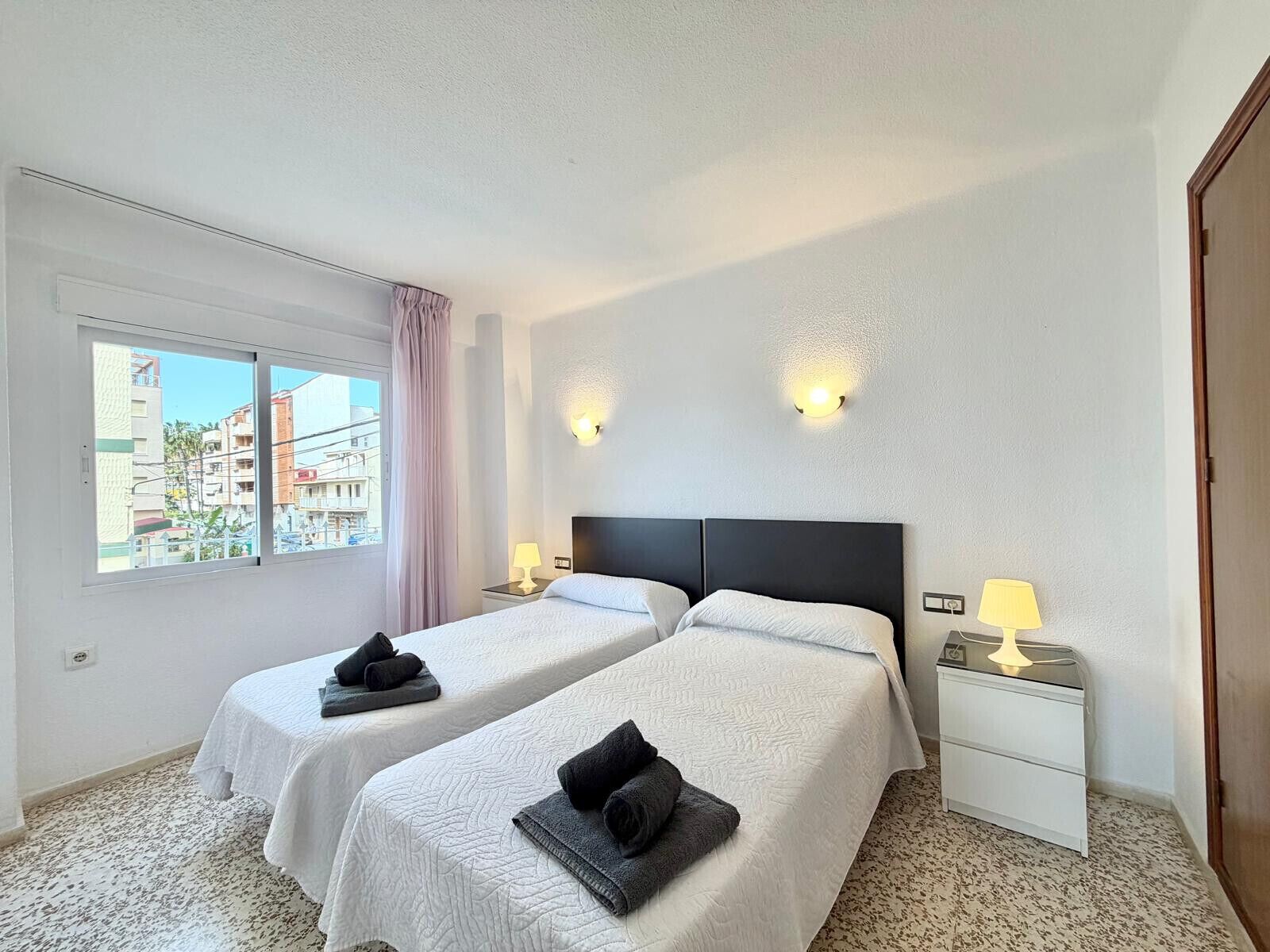 Une chambre d’hôtel avec deux lits, une fenêtre donnant sur des immeubles, une table de chevet avec une lampe et une porte.