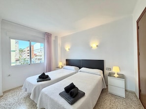 A hotel room with two beds, a window with a view of buildings, a nightstand with a lamp, and a door.