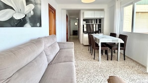 Ein modernes Wohnzimmer mit Sofa, Esstisch, Stühlen, einer Küchenzeile mit Schränken und einem großen Fenster.