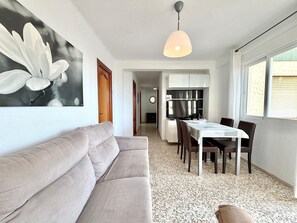 A modern living room with a sofa, dining table, chairs, a kitchen area with cabinets, and a large window.