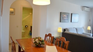 Un comedor con una mesa de madera, sillas y un florero. Se observa un sofá con cojines y una lámpara al fondo.