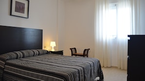 Una habitación de hotel con una cama grande, un escritorio con lámpara, una silla y un ventanal con cortinas.