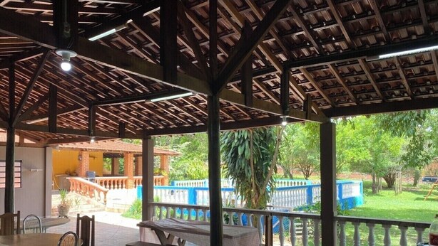 An outdoor patio area with wooden tables and chairs, a covered seating area, and a view of a garden with a white fence and greenery. - Chacara Zabotto (São Carlos)