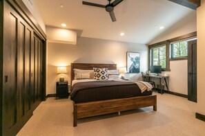 A bedroom with a wooden bed, a desk with a computer, a ceiling fan, and a window with a view of trees.